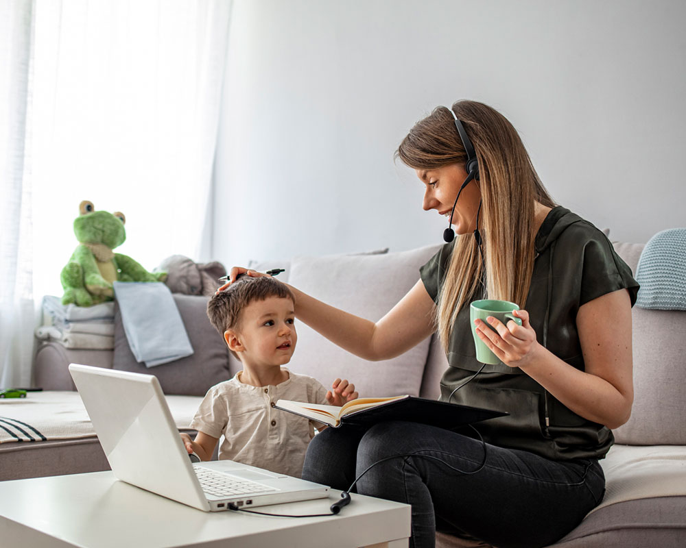 Woman holding coffee and sitting on the couch with her laptop while taking care of a young boy