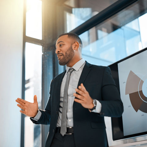 Man in a suit speaking with a chart on the screen behind him