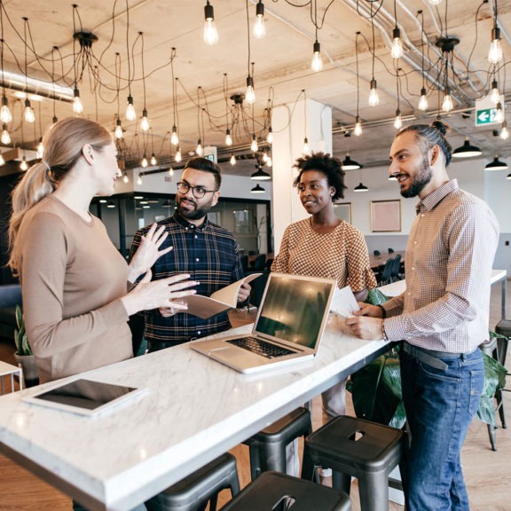 Four team members standing around a table and an open laptop talking