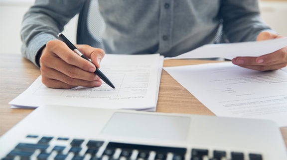 person writing with a pen and paper in front of a laptop