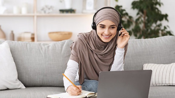 woman with headphones on writing in a notebook and looking at a laptop