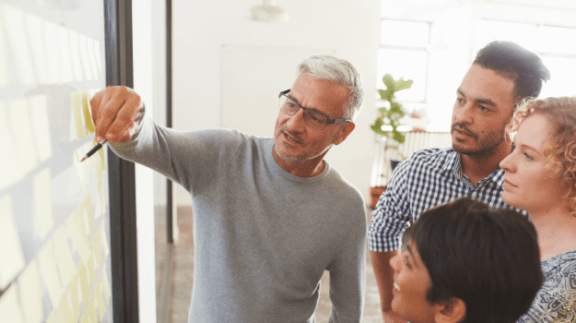 man with white hair pointing to a white board while others look on