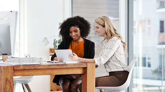 two women sitting at a wooden table looking at a laptop